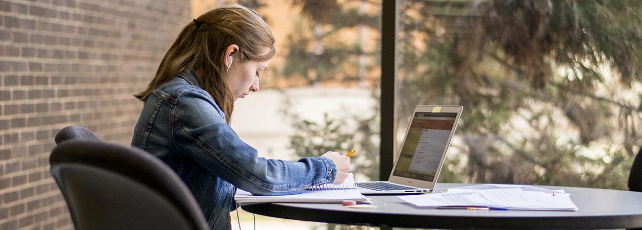 一名学生坐在奥利弗大厅走廊的桌子前，用她的笔记本电脑工作。